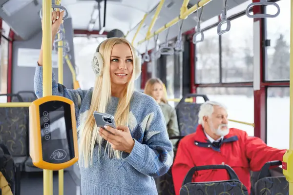 Front view of young blonde girl standing inside bus looking at window smiling holding handle. Happy female using phone in bus with blur old bearded man and girl on backround. Concept of city life.
