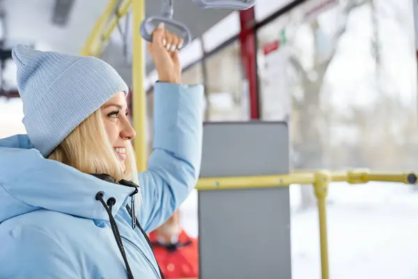 青いジャケットとキャップの笑顔の金髪の少女の側面の景色は 公共交通機関の窓を見ています バスに座っているぼやけの乗客が付いている女性の保持ハンドルの作物 冬の旅の概念 ストック画像