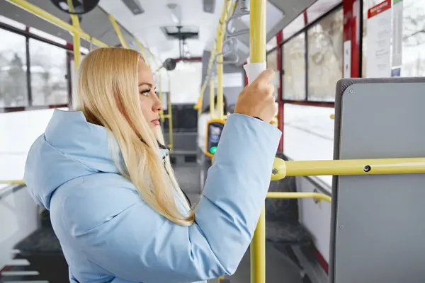移動輸送中に立っているナプキンを通して手すりを持っている青いジャケットを身に着けている美しい女性の側面 バスの窓を真剣に見つめている大人の女性の近く 輸送コンセプト ストック写真