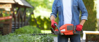 Arka bahçedeki çalıları şekillendirmek için elektrikli budayıcı kullanan koruyucu eldivenli isimsiz kaslı bir tamirci. Açık havada mevsimlik iş yaparken çit kesen erkek işçinin ekin manzarası. Bahçıvanlık kavramı.