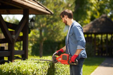 Koyu saçlı bahçıvan bahçedeki çit biçme makinesiyle fazla büyümüş çalıları kesiyor. Mevsimlik işler için modern bahçe malzemelerini kullanan, arka bahçedeki çitleri budayarak meşgul bir adamın yan görüntüsü. İş yeri kavramı.