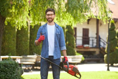 Bahçede elektrikli çit biçme makinesiyle dikilirken, eldivenli yakışıklı bahçıvan baş parmağını gösteriyor. Bitkileri budarken jest yapan mutlu esmer erkek işçinin portresi. İş kavramı.