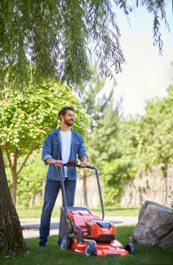 Sıradan giysiler içinde gülen sakallı adam bahçesinde modern çim biçme makinesiyle çimleri buduyor. Bahçede çim biçme makinesini iten, uzaktan bakan neşeli adamın ön görüntüsü. Bahçıvanlık kavramı.