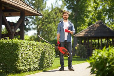 Arka bahçede elektrikli çit biçme makinesiyle dikilirken eldivenli, gülümseyen güçlü bahçıvan. Erkek peyzajcının, kutuları budarken jest yaptığı ön manzara. İş kavramı.