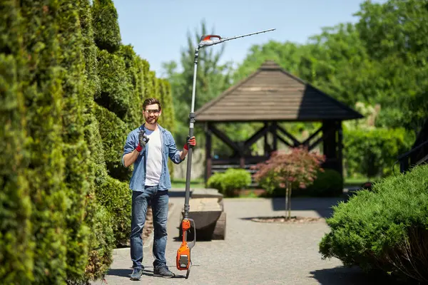 Dışarıda elektrikli çit biçme makinesiyle dururken mutlu erkek işçi iyi işareti gösteriyor. Kameraya bakarken çalı kesiciyle gülümseyen bahçıvanın ön görüntüsü. İş kavramı, hobi. 