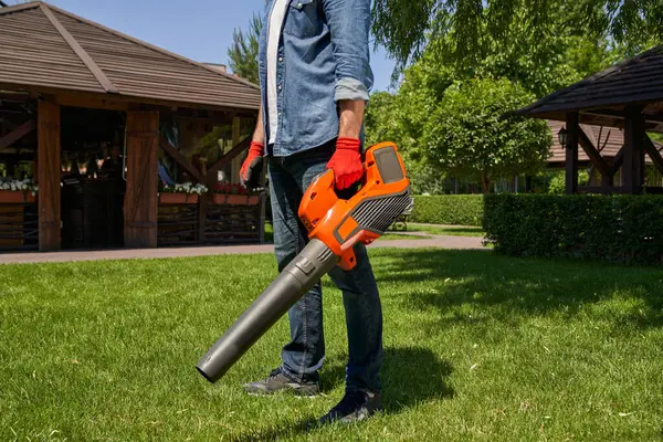 強力な男性庭師は 現代のガソリンコードレスリーフブロワーを使用しながら 裏庭庭の芝生を掃除しています 庭具で作業するデニム衣装の男の作物のビュー 季節的な仕事の概念 ストックフォト