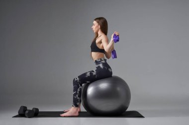 Aktif giyimli, lastik bant kullanan, kollarını arkadan uzatan genç bir kadın. Spor salonunda spor yapan bayan sporcunun yan görüntüsü, gri renkte izole edilmiş. Vücut bakımı, spor konsepti.