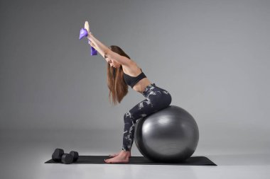 Gri pilates topunun üzerinde otururken lastik bant kullanan motive edici formda bir kadın. Kollarını kaldırmış kadın sporcu, formda top üzerinde çalışıyor, gri renkte izole edilmiş. Vücut bakımı, spor konsepti.