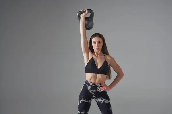 Strong Caucasian woman in sports bra, posing with grey kettle bell in studio. Portrait of strong long-haired female with raised arm, lifting kettle bell and looking at camera, isolated. Sport concept.