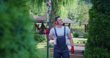 Strong man in overalls, safety glasses and gloves standing with electric trimmer in hands on back yard. Front view of handsome experienced gardener pruning green hedge at daytime. Concept of trimming.
