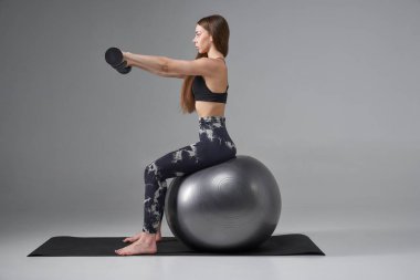 Spor sutyenli kararlı bir kadın, dambıl kullanıyor, kollarını kaldırıyor, antrenman yapıyor. Kol halterleriyle spor yapan kadın sporcu, gri renkte izole edilmiş. Vücut bakımı, spor konsepti.