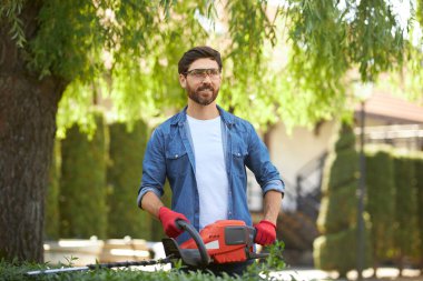 Mutlu yakışıklı bahçıvan güneşli bir günde çim biçme makinesiyle çalı çırpı kesiyor. Bahçıvanlık ekipmanlarıyla çalışan sakallı tamircinin portresi. Mevsimlik iş kavramı.