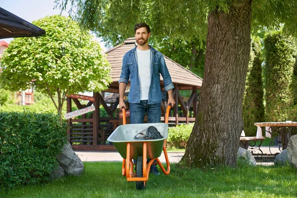 화창한 정원에서 휠바로를 카메라를 보면서 정원을위한 카트를 사용하여 셔츠에 사람의 스톡 이미지