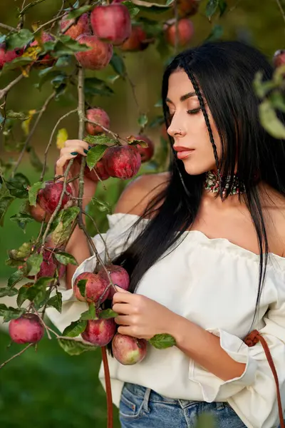Sad, dark-haired woman in shoulderless white shirt, holding tree branches, laden with red apples. Close up of thoughtful pierced girl posing in apple orchard with closed eyes. Harvest, beauty concept.