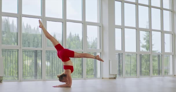 高级女性瑜伽选手身穿红色衣服 在宽敞的画室里倒立着瑜伽姿势 从侧面看白种人女人的发髻慢动作练习瑜伽 健身观念 — 图库视频影像
