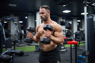 Spor salonunda çalışan kararlı, yakışıklı bir erkek halterci. Saç stili olan sakallı bir adamın portresi, dambılları kaldırıyor ve bulanık arka plana bakıyor. Vücut geliştirme, spor konsepti.