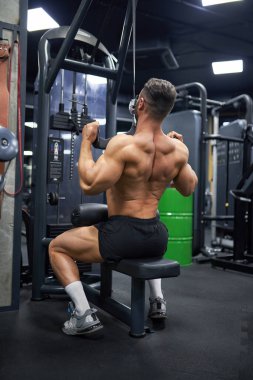 Güçlü, kaslı bir adam spor salonunda egzersiz yaparken antrenman aparatını çekiyor. Şortlu, sırt kası geliştiren, spor yapan kimliği belirsiz sağlıklı bir adamın arka planı. Spor, yaşam tarzı, vücut geliştirme konsepti..