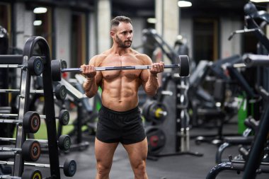 Azimli, güçlü bir atlet spor salonunda ağır halter tutuyor. Kaslı beyaz adamın portresi halteri kaldırıyor, kasları gergin tutuyor, arka planda bulanıklık var. Vücut geliştirme, halter konsepti.