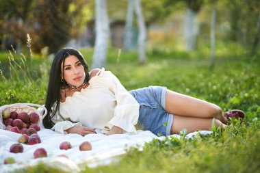 Kaygısız koyu saçlı güzellik güneşli bir günde dağınık elmalarla çevrili yerde dinleniyor. Sonbahar bahçesinde beyaz halıda uzanan mutlu genç bayanın yan görüntüsü. Piknik, güzellik kavramı. 