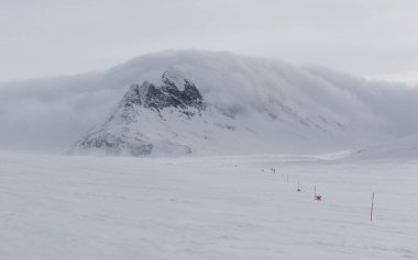 Karda yarı gömülü Nordic kayak turu işaretleri Sylarna dağlarında yemini gösteriyor. Arka plandaki fırtına bulutları dağın üzerinden yuvarlanıyor..