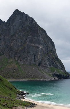 Kvalvika sahilinin beyaz kumlarını gösteren bir fotoğraf, turkuaz mavi su ve engebeli bir dağ zeminiyle. Kıyıda küçük bir çadır görülebilir..