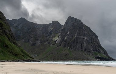 Norveç, Lofoten 'deki Kvalvika plajının arkasındaki karanlık dağlarda bir güneş ışığı parlıyor.