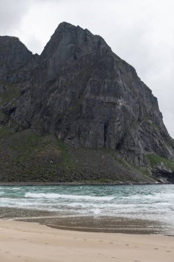 Kvalvika plajı karanlık, engebeli dağların ve Norveç 'te Lofoten' de bulutlu bir gökyüzünün altında yer alır.