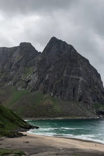 stock image Stormy skies and rugged cliffs rise above Kvalvika beach in Lofoten, Norway. A solitary person walks along the sand.