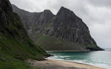 Lofoten, Norveç 'teki Kvalvika plajı bulutlu bir günde. Kıyı dağları, okyanus ve kumlu bir plajda çekilmiş bir manzara fotoğrafı..