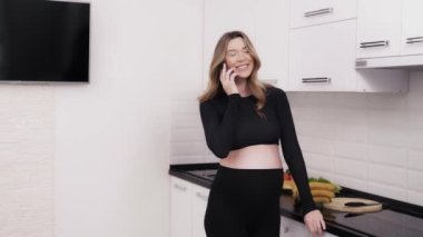 Hamile kadın yemek pişirmeden önce telefonla konuşuyor. Hoş sohbetler onu mutlu ediyor. Gelecekteki anne cep telefonunda konuşurken karnına dokunuyor. 