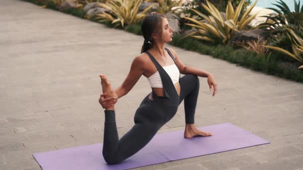 中庭の庭でヨガを練習柔軟な体を持つかなり若い女性 女性は太腿とふくらはぎを伸ばすために運動する バランスを改善し ストレスを軽減し 心を静めるための物理的な活動 — ストック動画