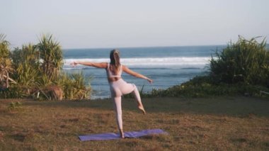 Sıska bir kızın bir bacağı havada yoga yaparken görüntüsü. Kadın vücut dengesini geliştirmek için egzersiz yapıyor. Sağlık ve zindelik. Deniz manzarasına karşı antrenman 