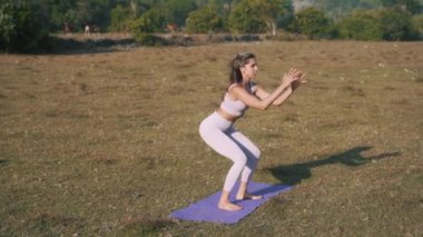 Genç bir kadın sahilde antrenman yapıyor. Bacaklarınızı çalıştırmak için vücudunuzun alt kısımlarını çalıştırın. Çömelip denize bakan bir kız. Sporla sağlıklı bir yaşam tarzı, uzun mesafeli çekim. 