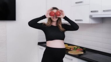 Happy pregnant woman taking joy in playing and dancing with tomatoes in the kitchen. Future mother enjoying the time of baby expectation and going to cook healthy meals