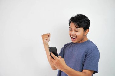 Heyecanlı gülümseme ya da donanma tişörtlü mutlu Asyalı genç adam beyaz arka planda izole edilmiş bir şekilde telefon tutuyor.