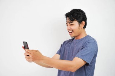 Heyecanlı gülümseme ya da donanma tişörtlü mutlu Asyalı genç adam beyaz arka planda izole edilmiş bir şekilde telefon tutuyor.