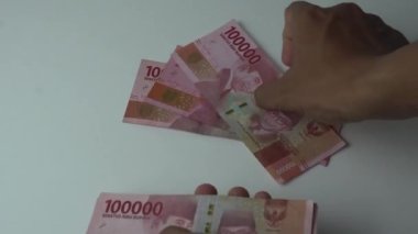 Adamın eli, yüz bin rupia banknotunu sayıyor. Beyaz arka planda Endonezya parası. Beyaz bir masaya yatarak para saymak.