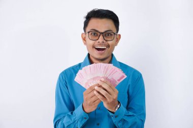 Endonezya taraftarı mavi gömlekli ve gözlüklü yakışıklı genç Asyalı iş adamının beyaz arka planda izole edilmiş ellerinde banknotlar tutmasına şaşırmış ya da şok olmuştu.