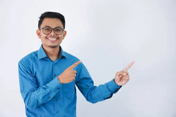 白い背景に隔離された青いシャツを身に着けている側面を指差すポーズ指で笑顔のメガネを持つハンサムなアジア系男性従業員 — ストック写真