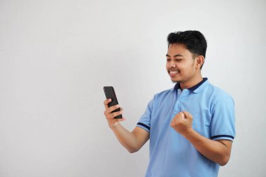 Heyecanlı ya da mutlu Asyalı genç bir adam telefon tutuyor. Yumruklarını sıkmış, mavi polo tişört giyiyor. Beyaz arka planda izole edilmiş.