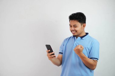 Heyecanlı ya da mutlu Asyalı genç bir adam telefon tutuyor. Yumruklarını sıkmış, mavi polo tişört giyiyor. Beyaz arka planda izole edilmiş.