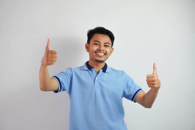 Mavi tişörtlü neşeli Asyalı adam portresi beyaz arka planda izole edilmiş kameraya gülümsüyor ve başparmaklarını gösteriyor.
