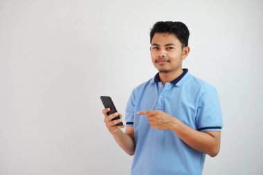 Gülümseyen ya da mutlu Asyalı bir adam mavi tişört giymiş elinde telefonla akıllı telefonu işaret ediyor, beyaz arka planda izole edilmiş.