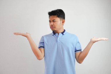 ciddi yüzlü Asyalı bir adam iki kolu da açık, yanında beyaz arka planda mavi tişört giyiyor.