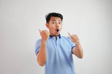 Şok olmuş Asyalı bir adam, ağzı açık, parmakları arkasını işaret eden beyaz arka planda mavi tişört giyen.