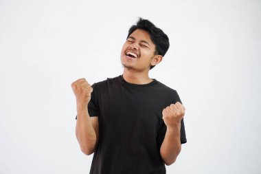 Kendine güveni tam, yakışıklı Asyalı adam siyah tişört giyerek el kaldırıyor ve yumruklarını sıkıyor. Mutluluğu hissetmek, beyaz arka planda izole edilmiş yumruğu başarıyı gösterir.