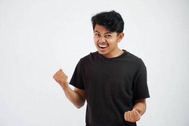 Kendine güveni tam, yakışıklı Asyalı adam siyah tişört giyerek el kaldırıyor ve yumruklarını sıkıyor. Mutluluğu hissetmek, beyaz arka planda izole edilmiş yumruğu başarıyı gösterir.