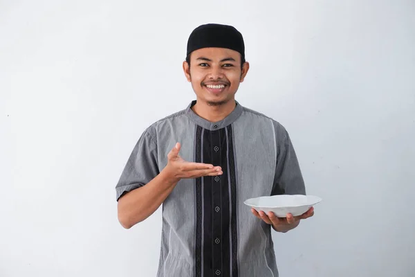白地にグレーのイスラム教徒の服を着て空のディナープレートを持ちながら 興奮した表情を示す笑顔や幸せな若いアジアのイスラム教徒の男 — ストック写真