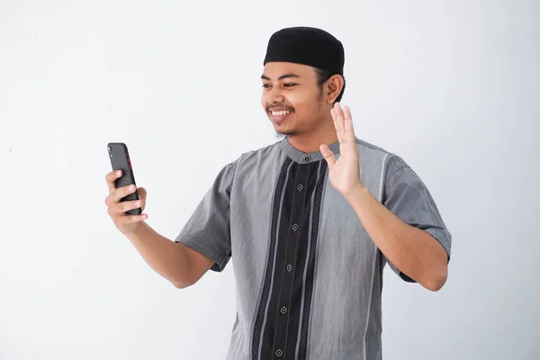 ココ姿のハンサムなアジア系ムスリム男性の肖像画や 白い背景に隔離された携帯電話でビデオ通話や笑顔をするスマートフォンを使って — ストック写真