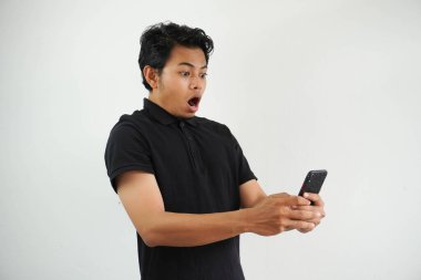 Polo tişört giyen genç Asyalı şok olmuş bir adam telefonuna sürpriz bir ifadeyle bakarken, mobil cihazlar inanılmaz haberler öğrenirken, reklam satışından hayrete düşerken beyaz arka planda izole edilmiş.